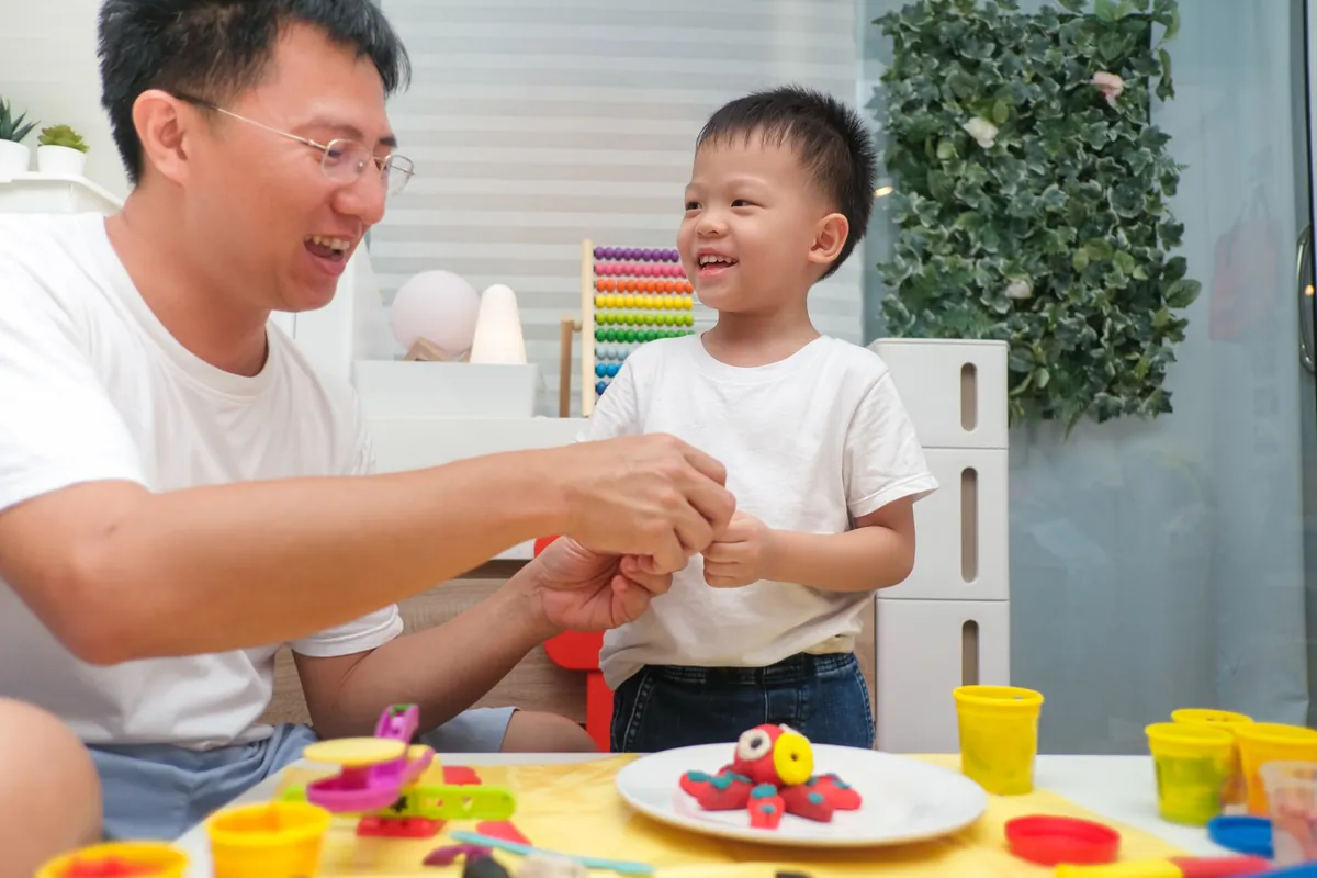 A father and his young son laughing together at a craft table covered in modeling clay