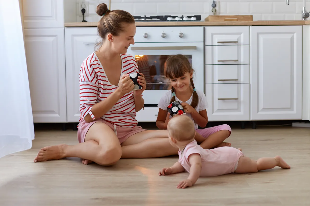 A mother and her young daughter smiling at each other on a kitchen floor while the baby crawls beside them