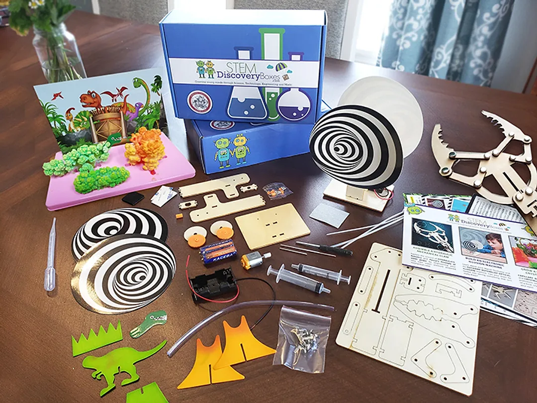 Two royal-blue STEM Discovery Boxes on a dining table with a month's contents laid out around them: a finished dinosaur crystal garden, a spiral optical-illusion spinner on its stand, a wooden hydraulic claw, laser-cut wood sheets, syringes and tubing, a battery pack and motor, and the printed project cards