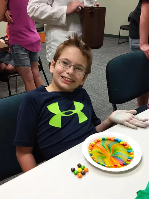 A smiling boy at a table beside a plate of candies bleeding their colours into a rainbow