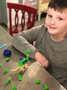 A smiling boy at a table next to a dinosaur model made of green and blue modeling clay, with more clay pieces scattered on the table