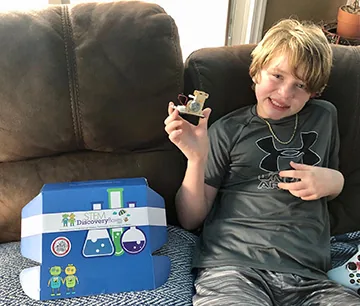 A boy sitting on a couch holding up a small wooden build with a wire attached, next to a STEM Discovery Box