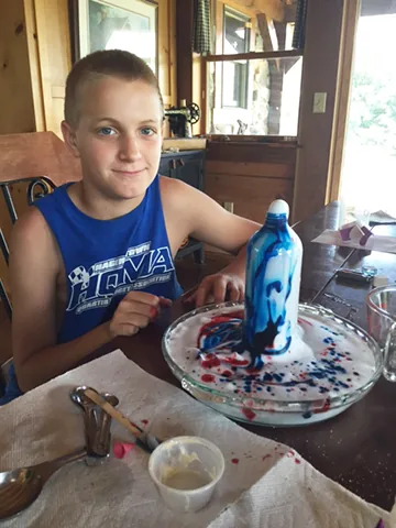 A boy at a table smiling beside a science experiment with blue liquid foaming out of a bottle onto a plate