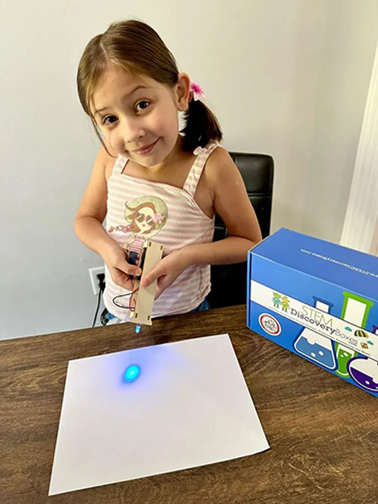 A girl at a table holding up her finished build beside a blue STEM Discovery Box, a small light glowing on the paper in front of her