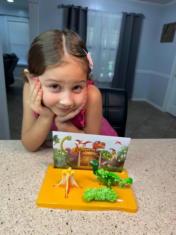 A smiling girl resting her chin on her hands behind a dinosaur diorama she made