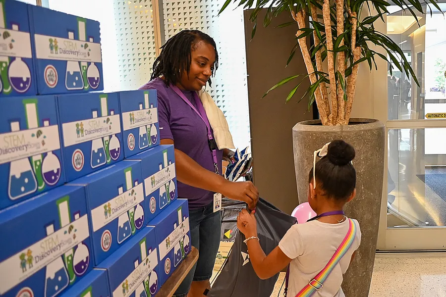 A woman handing a STEM Discovery Box to a girl at an event, with a wall of stacked boxes behind them