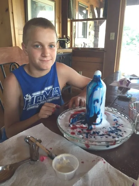 A boy at a table smiling beside a bottle with blue foam pouring out of it