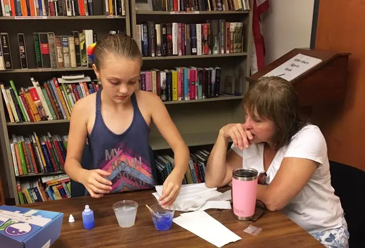 A girl and a woman working on a project together at a table in front of library bookshelves