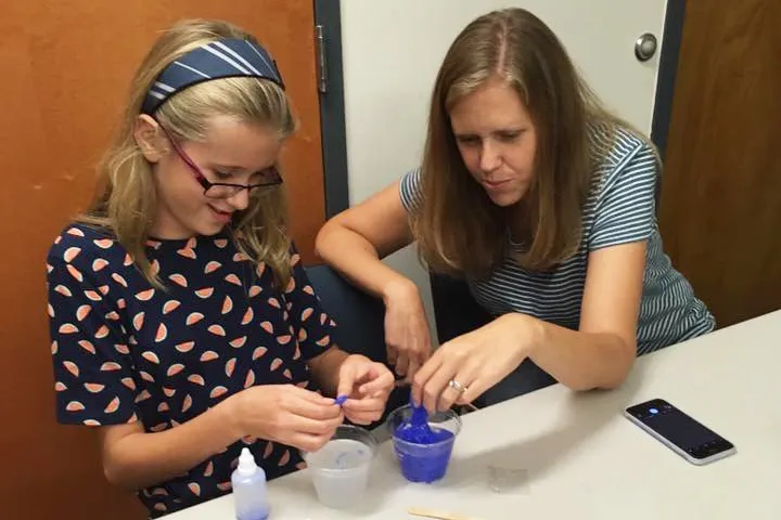 A girl and a woman at a table mixing blue slime in a cup