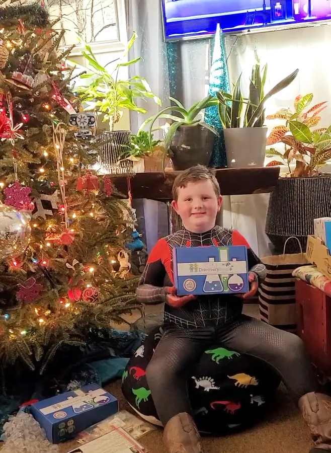A boy sitting by a Christmas tree holding up a STEM Discovery Box