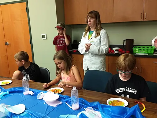 Four children around a table with paper plates of candies bleeding color into water, while an adult in a white lab coat stands and leads the activity