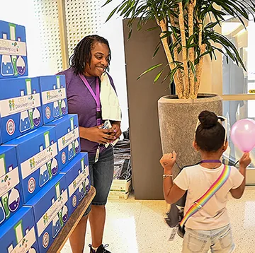 A woman standing beside a stack of STEM Discovery Boxes smiles down at a young girl holding a pink balloon
