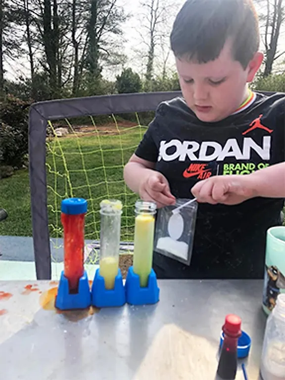 A boy outdoors opening a supply packet beside three tall tubes of coloured liquid in a blue stand