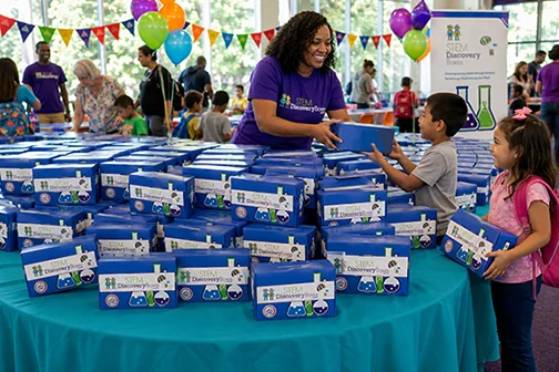 A woman in a purple STEM Discovery Boxes T-shirt hands a blue STEM Discovery Box to a boy across a table stacked with boxes, while a girl beside him holds her own box, at an indoor event with balloons