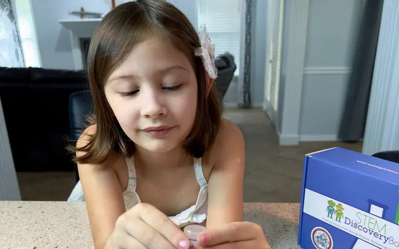 A girl working through a hands-on STEM Discovery Box project at her kitchen counter at home, with the blue box beside her