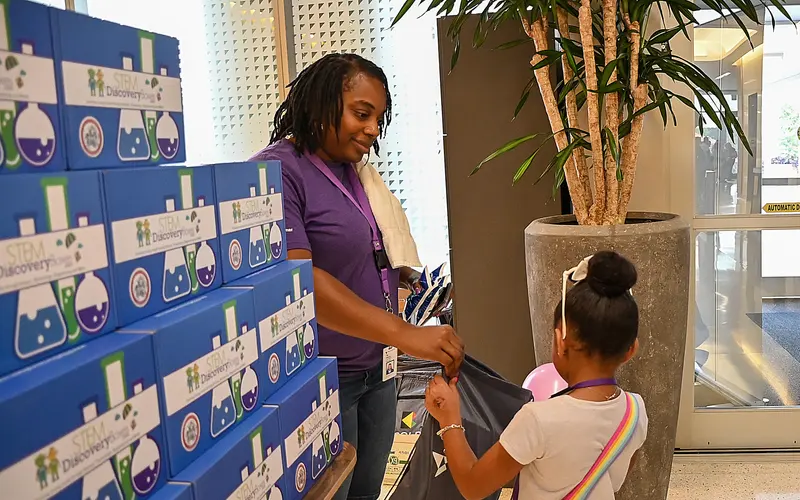 A STEM Discovery Boxes team member handing a kit to a child at a community STEM event, in front of a stacked wall of blue boxes