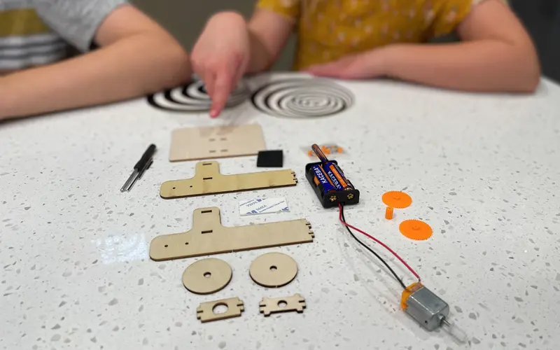 Two children at a counter with the laser-cut wooden parts, motor, battery pack and optical spiral discs from a STEM Discovery Box laid out in front of them