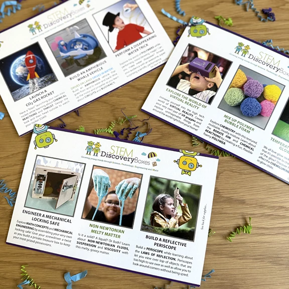 Three printed STEM Discovery Boxes lesson cards fanned on a wooden table, each showing photo instructions and the science behind a project