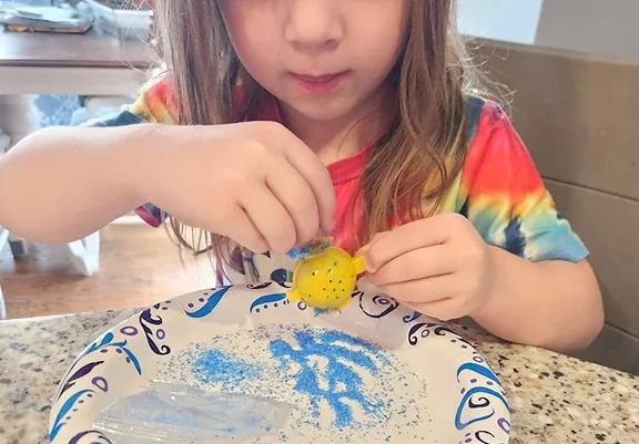 A girl holding the yellow bouncy ball she made over a plate of leftover crystals