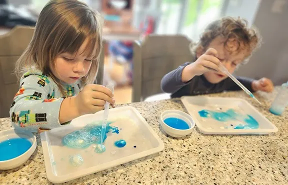 Two small children using pipettes over trays of blue liquid at a table