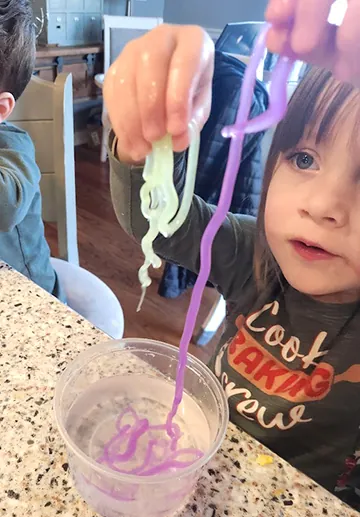 A young girl lifting purple and green polymer worms out of a cup with her fingers, her little brother beside her