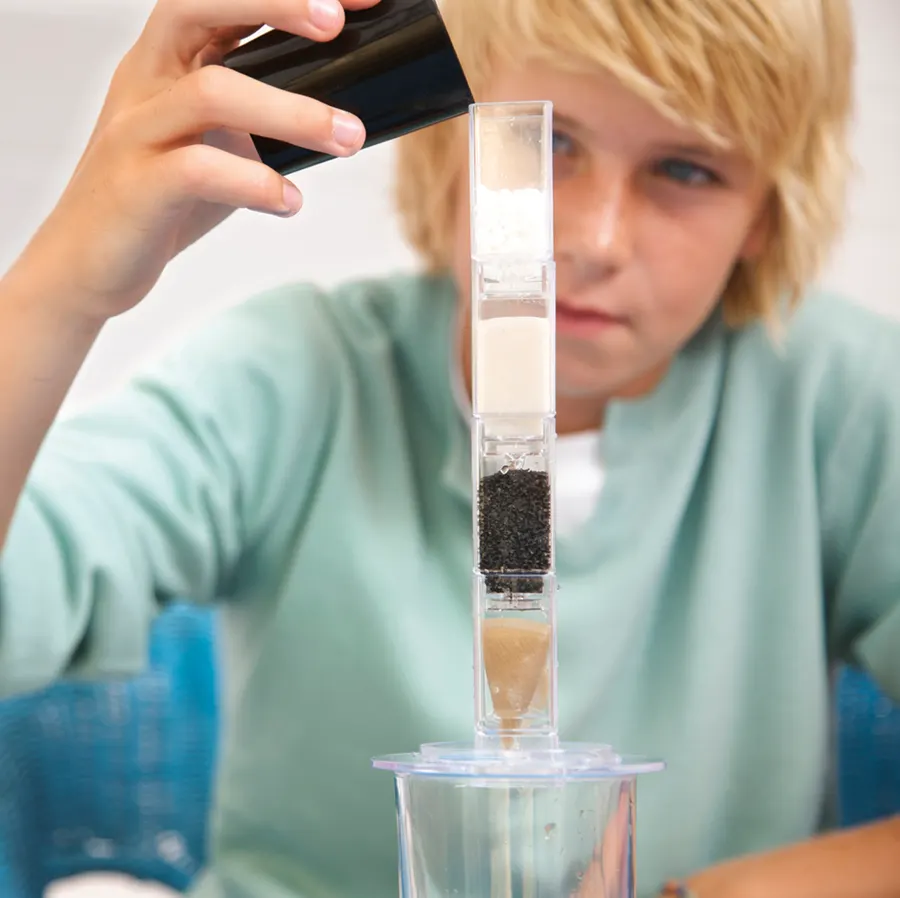 A boy pouring dirty water into a clear filtration column layered with sand, carbon and granite