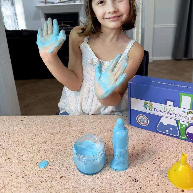 A girl holding up both hands covered in blue foam, with the foam still erupting from a jar and a bottle on the counter beside her STEM Discovery Boxes kit