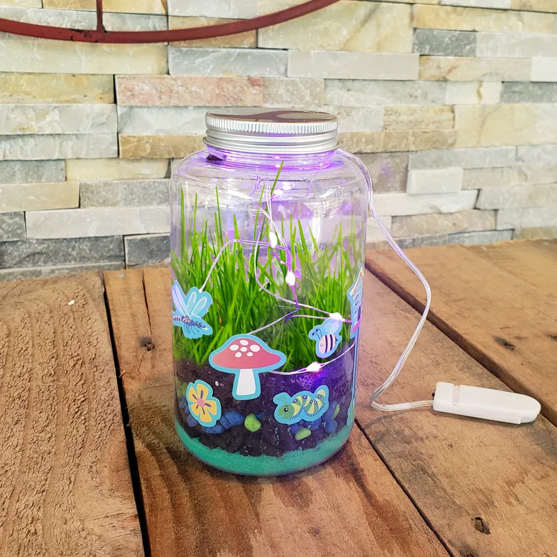 A lit terrarium jar on a wooden table: grass growing above layers of colored sand and stones, purple string lights inside and garden stickers on the glass