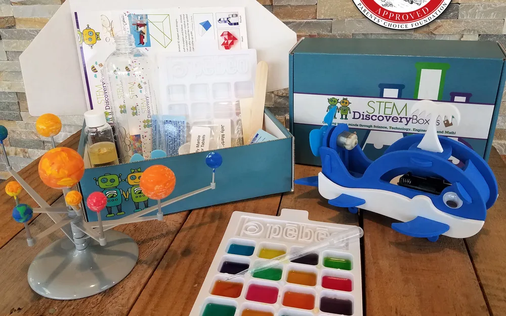 An open STEM Discovery Box beside a hand-built solar system model, a motorized whale, and a watercolor paint tray